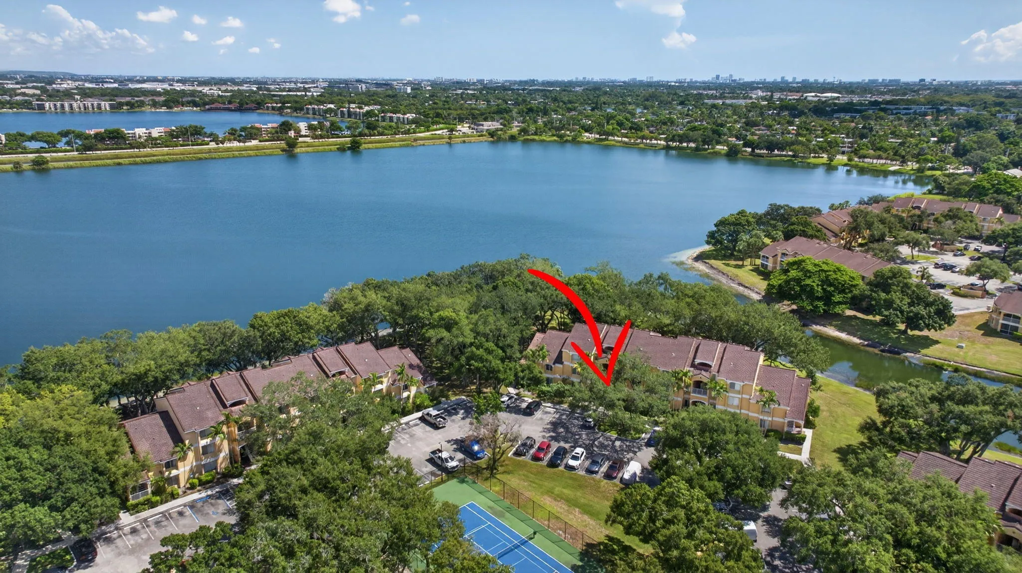 Waterfront multi-story building featuring a tile roof, surrounded by mature trees, with a surface parking lot and a tennis court