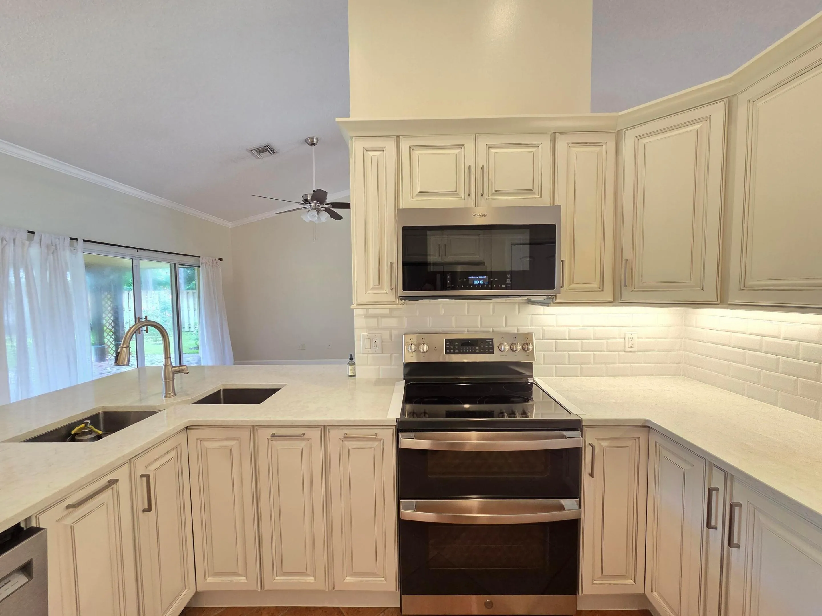 U-shaped kitchen featuring cream-colored cabinetry, stainless steel appliances, dual undermount sinks, white subway tile backsplash, and light-toned countertops