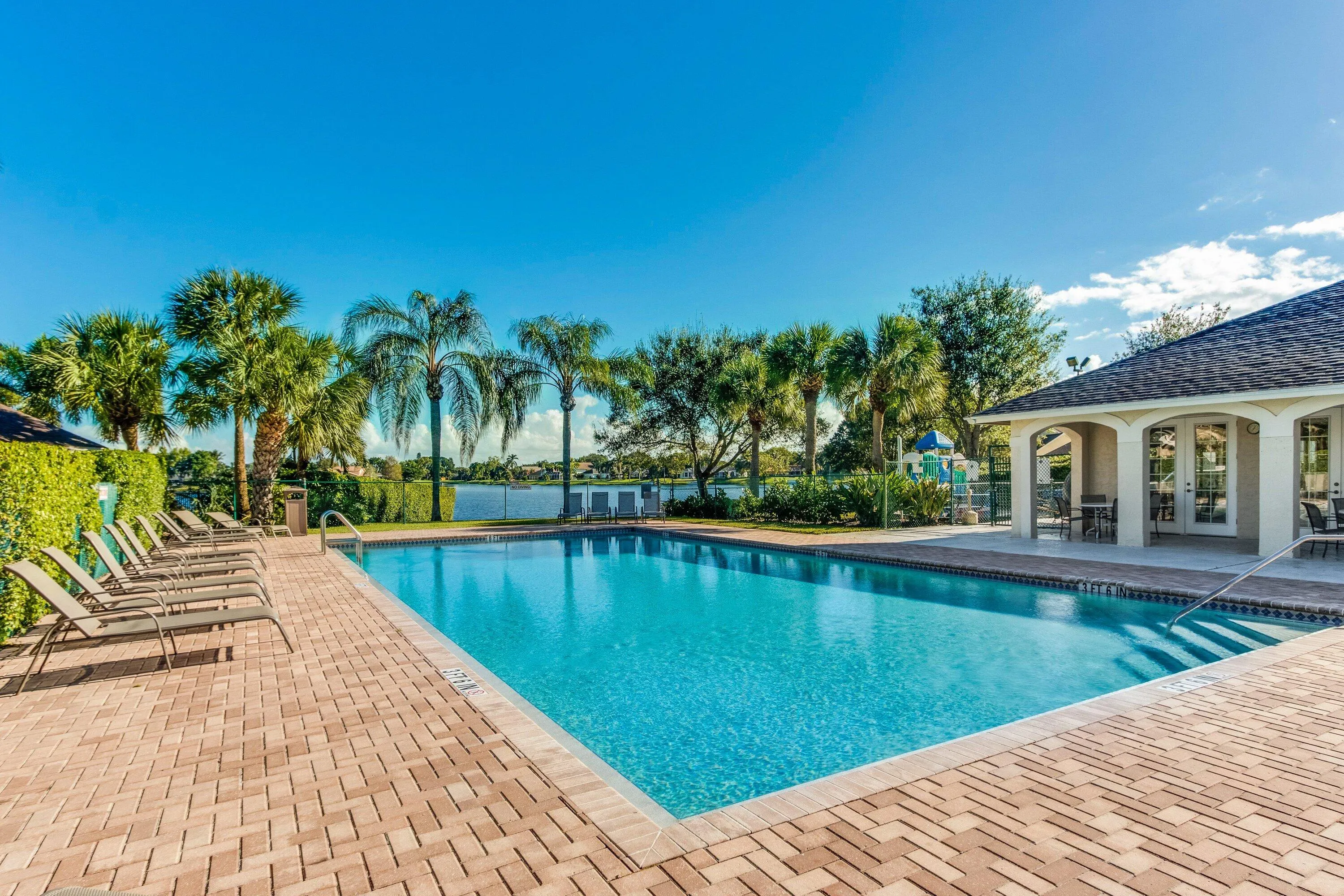 Expansive swimming pool with integrated steps, surrounded by a brick paver deck, a pool house with arched entries, and mature palm trees