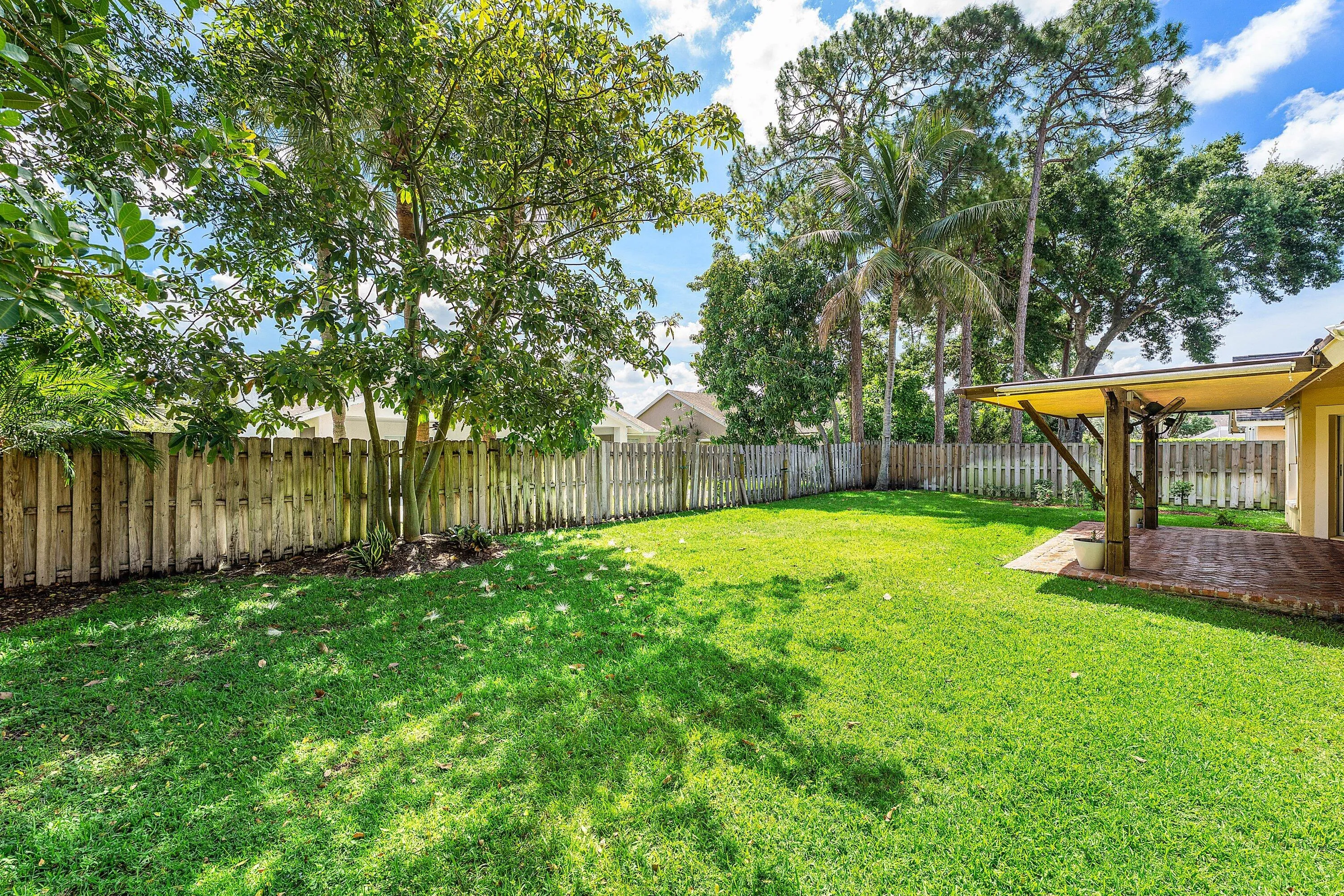 Expansive grassy yard featuring a fence, mature trees, and a covered brick patio