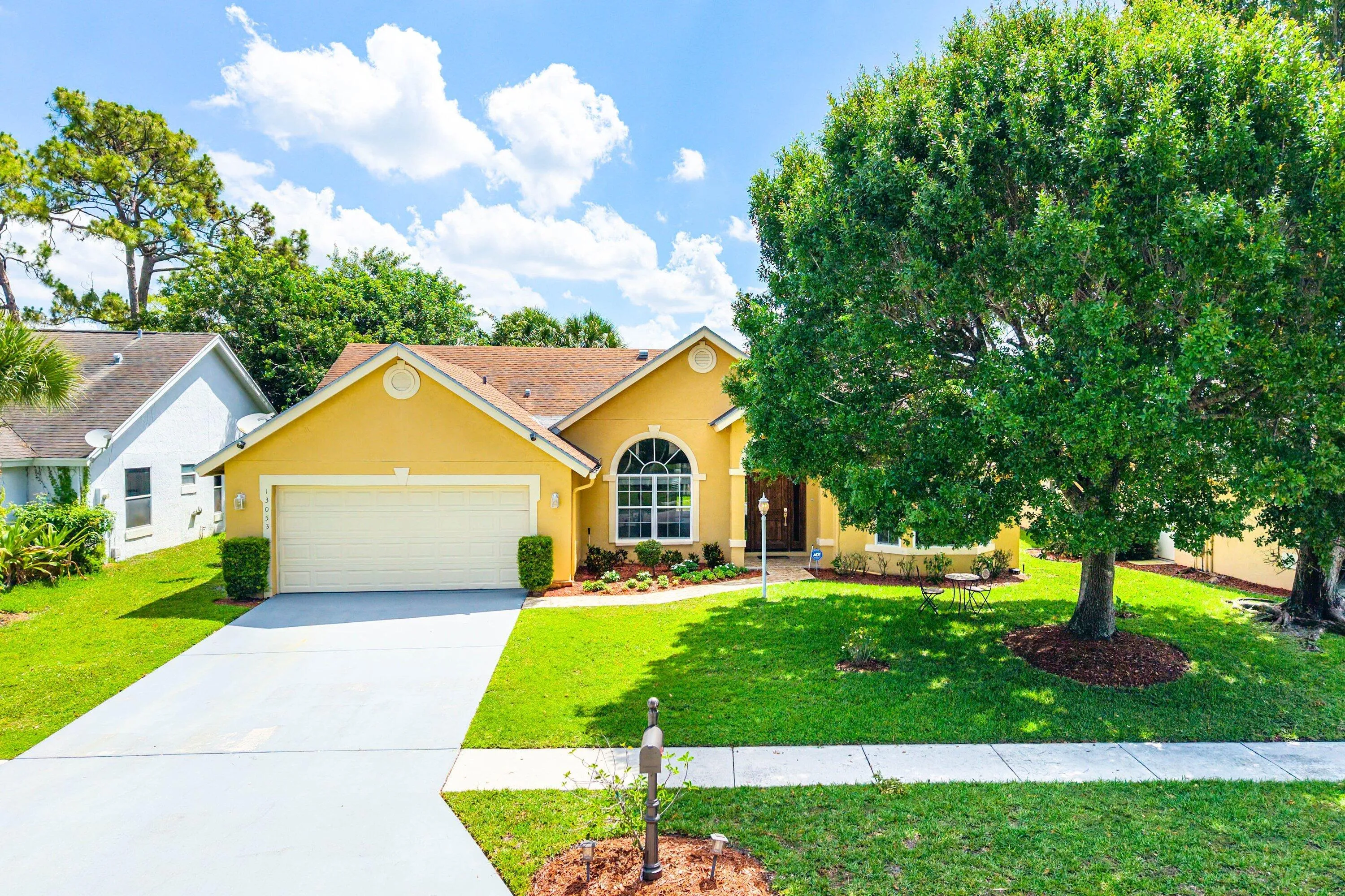 Warm yellow exterior with a prominent arched window, attached two-car garage, manicured front lawn, and mature shade trees