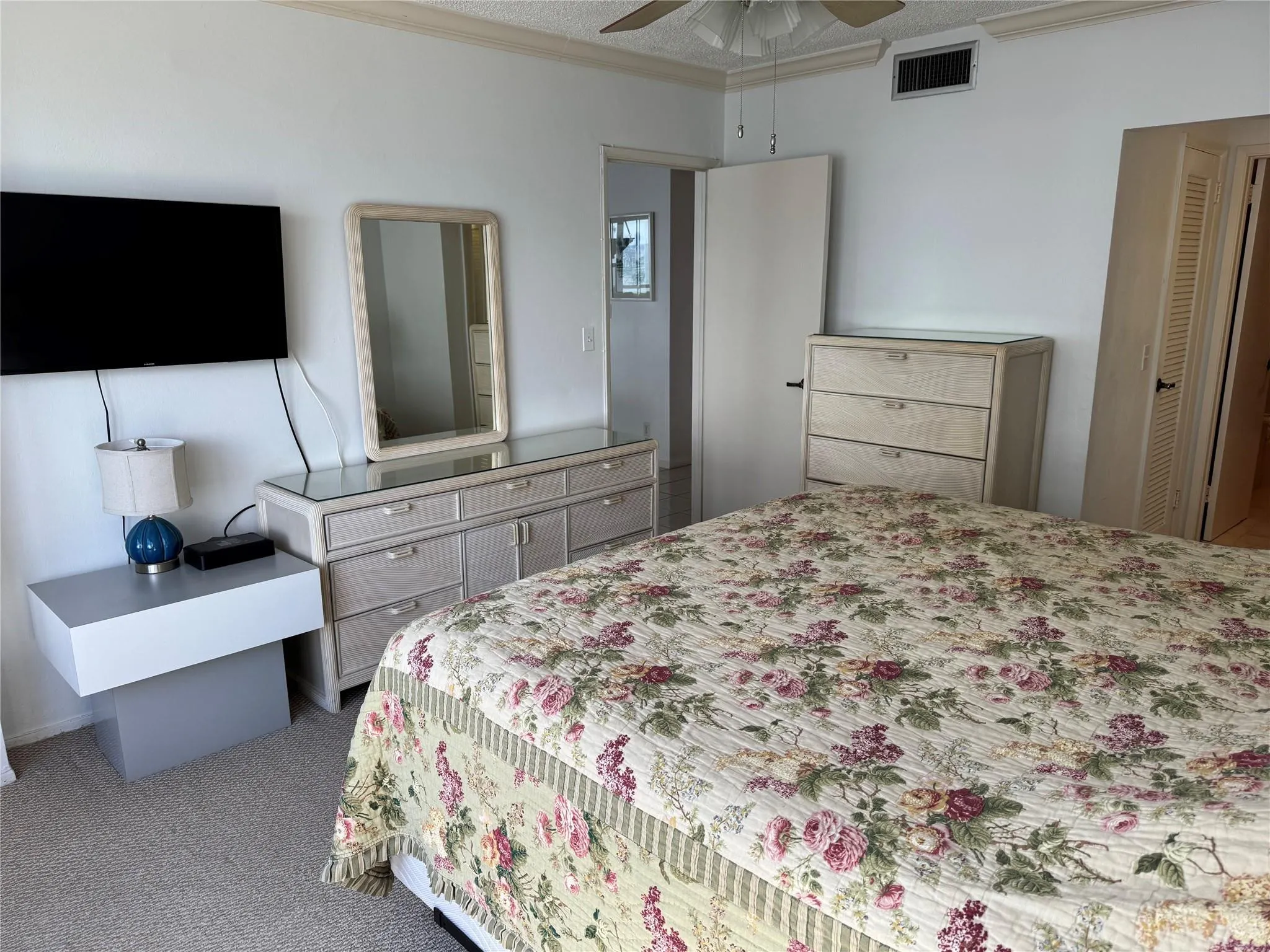 Spacious primary bedroom suite featuring dual mirrored dressers with wood-finish cabinetry, a coordinating wardrobe armoire, wall-mounted flat-screen television, ceiling fan, and carpet flooring throughout