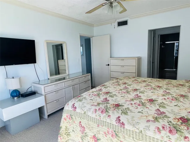 Spacious primary bedroom featuring carpeted flooring, ceiling fan with integrated lighting, mirrored wardrobe closet, wood-finish dresser with attached mirror, flat-screen television mount, and direct access to an ensuite bathroom