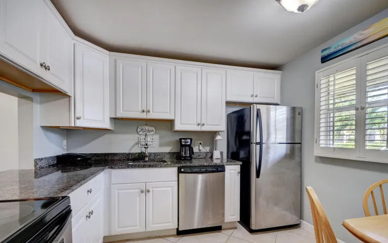 White cabinetry, dark stone countertops, stainless steel appliances, tile flooring, and a window with plantation shutters