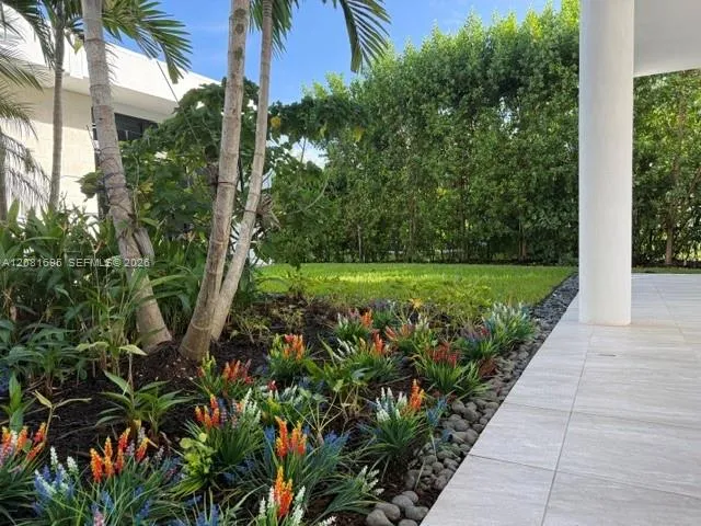 Covered patio with large-format tile flooring and a structural column, complemented by a landscaped garden featuring mature palms, tiered shrubbery, and river rock borders
