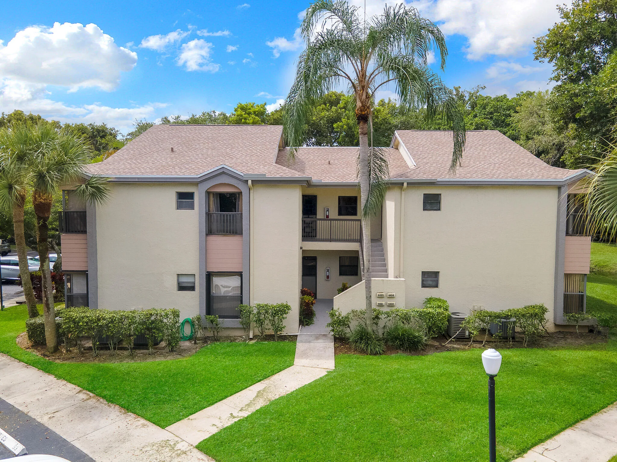 Two-story stucco exterior with a multi-gable roof, mature palm trees, manicured shrubbery, and a concrete walkway