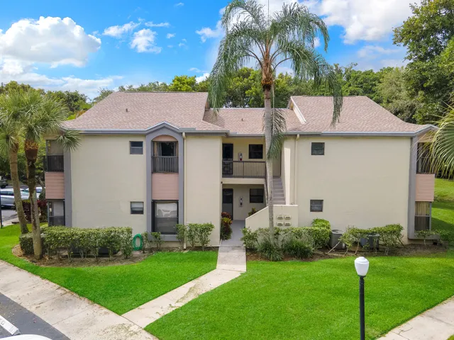 Two-story stucco exterior with a multi-gable roof, mature palm trees, manicured shrubbery, and a concrete walkway