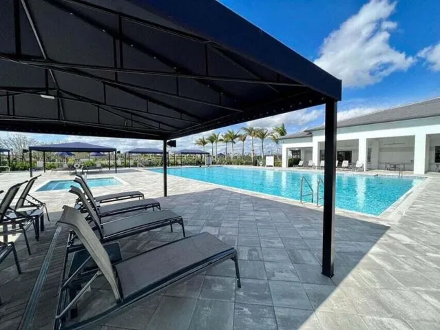 Community pool area featuring a large swimming pool, separate spa, shaded lounge seating, and expansive paver decking