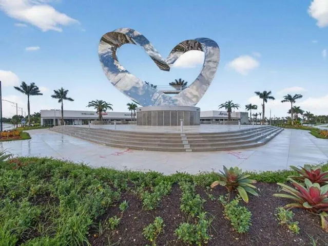 Expansive plaza with a prominent metallic sculpture, tiered concrete seating, mature palm trees, and lush landscaping