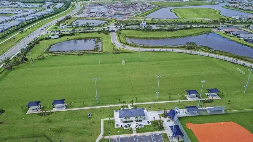 Expansive green field with multiple sports amenities including a baseball diamond, soccer goals, covered seating areas, and a central clubhouse structure