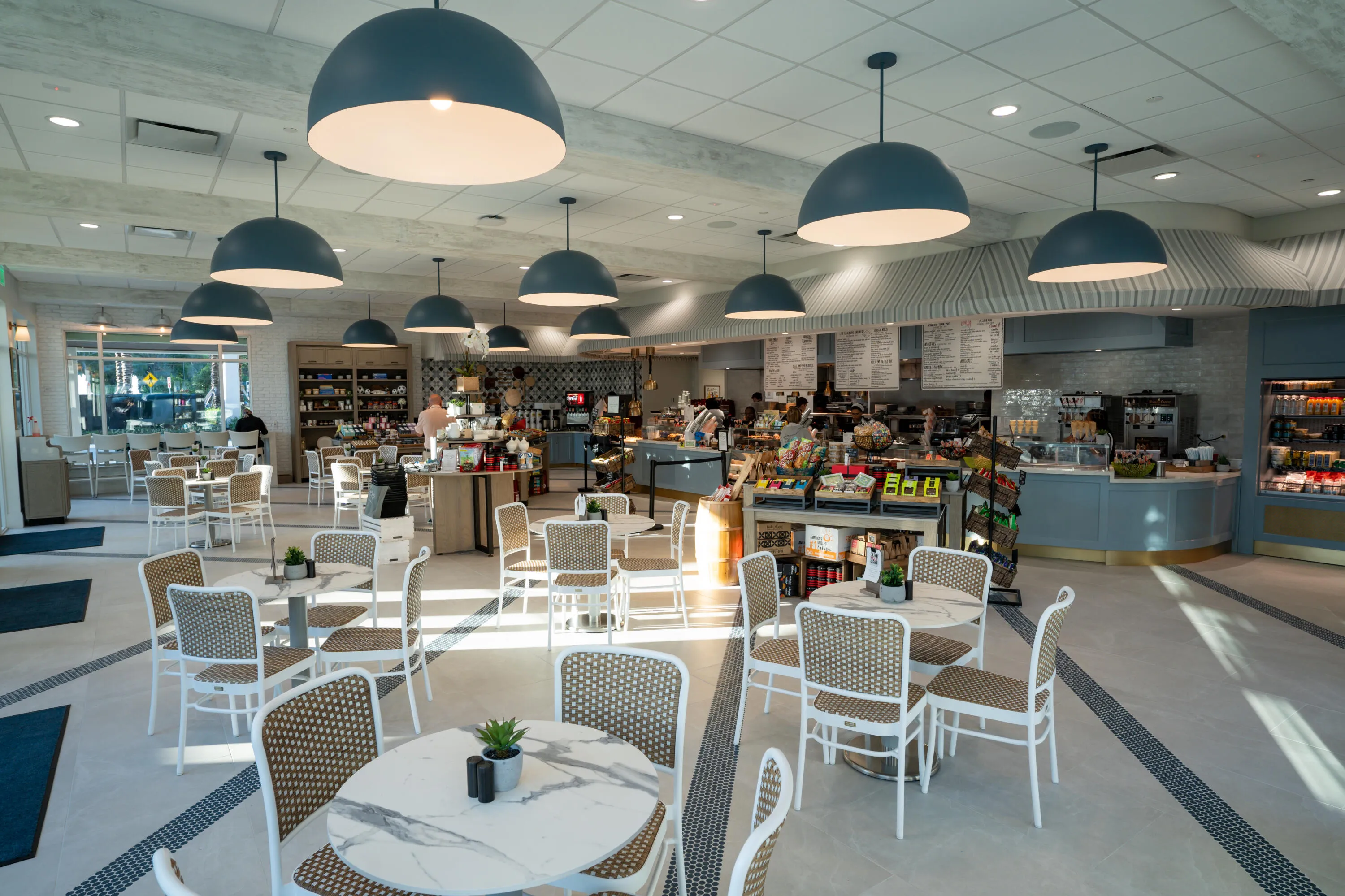 Modern commercial dining space featuring multiple large dome pendant fixtures with integrated lighting, polished concrete flooring, white mesh-back seating, and an open layout with integrated service counters and display shelving throughout the venue
