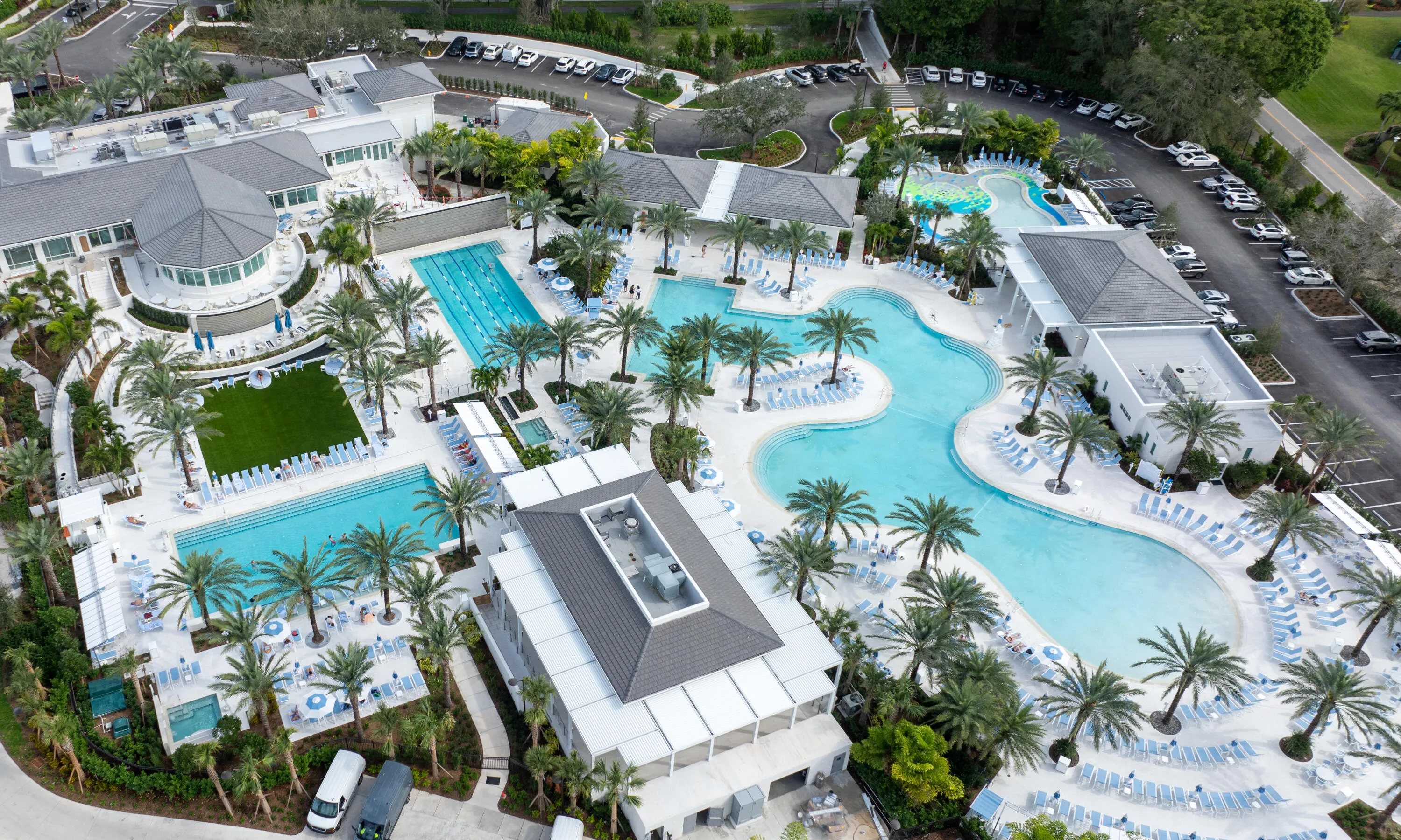 Expansive resort-style pool complex featuring multiple swimming pools, mature palm trees, various lounge areas, and surrounding structures with gray shingle roofs