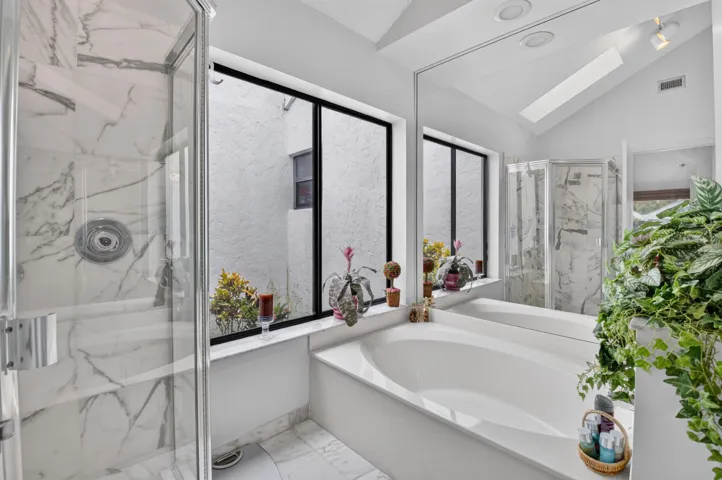 Recessed tub with an expansive mirror, enclosed shower with marble-look tile, and a vaulted ceiling with a skylight