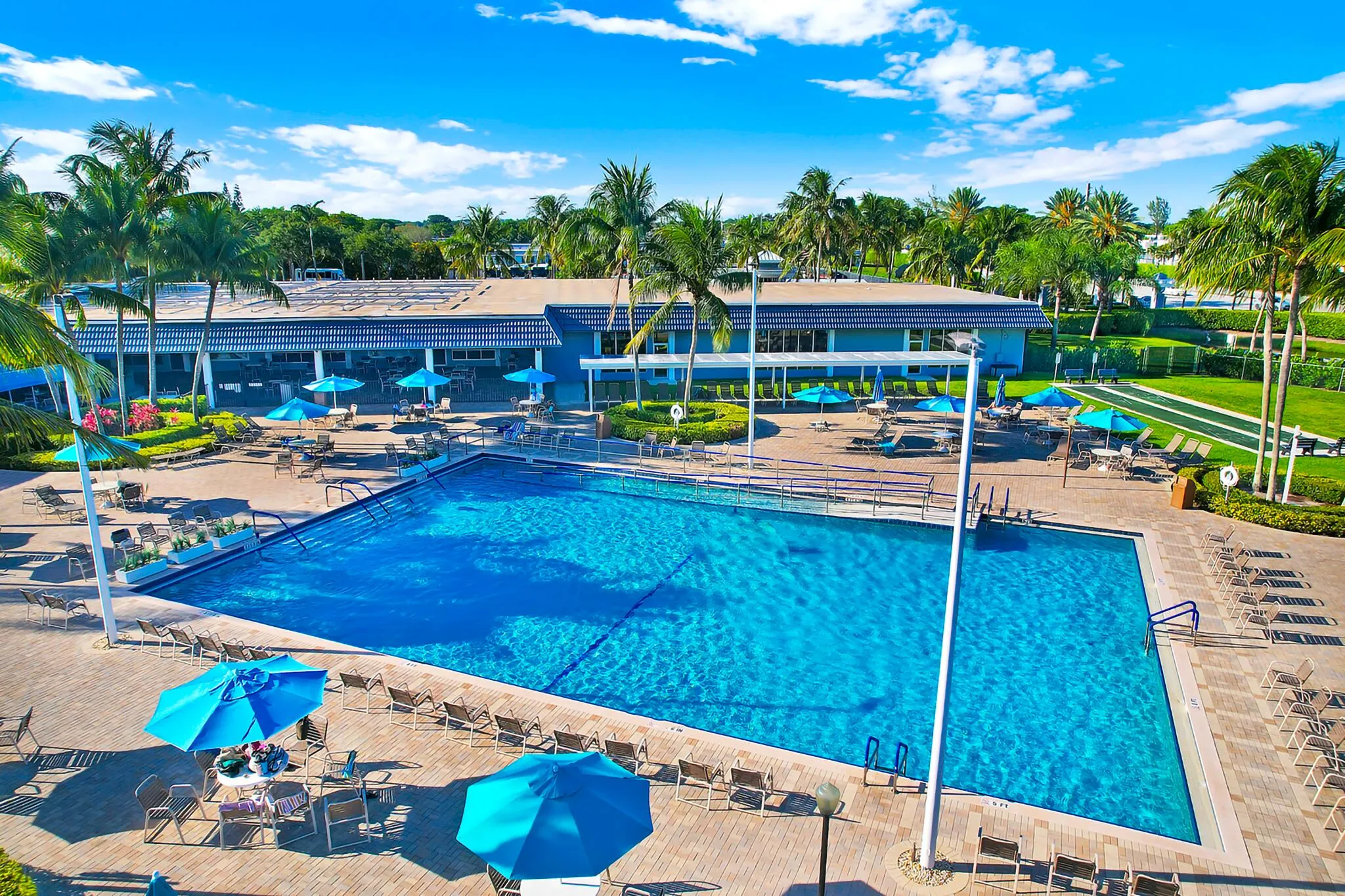Expansive outdoor swimming pool with surrounding patio space, featuring a building with a covered walkway, a large lawn area, and mature palm trees