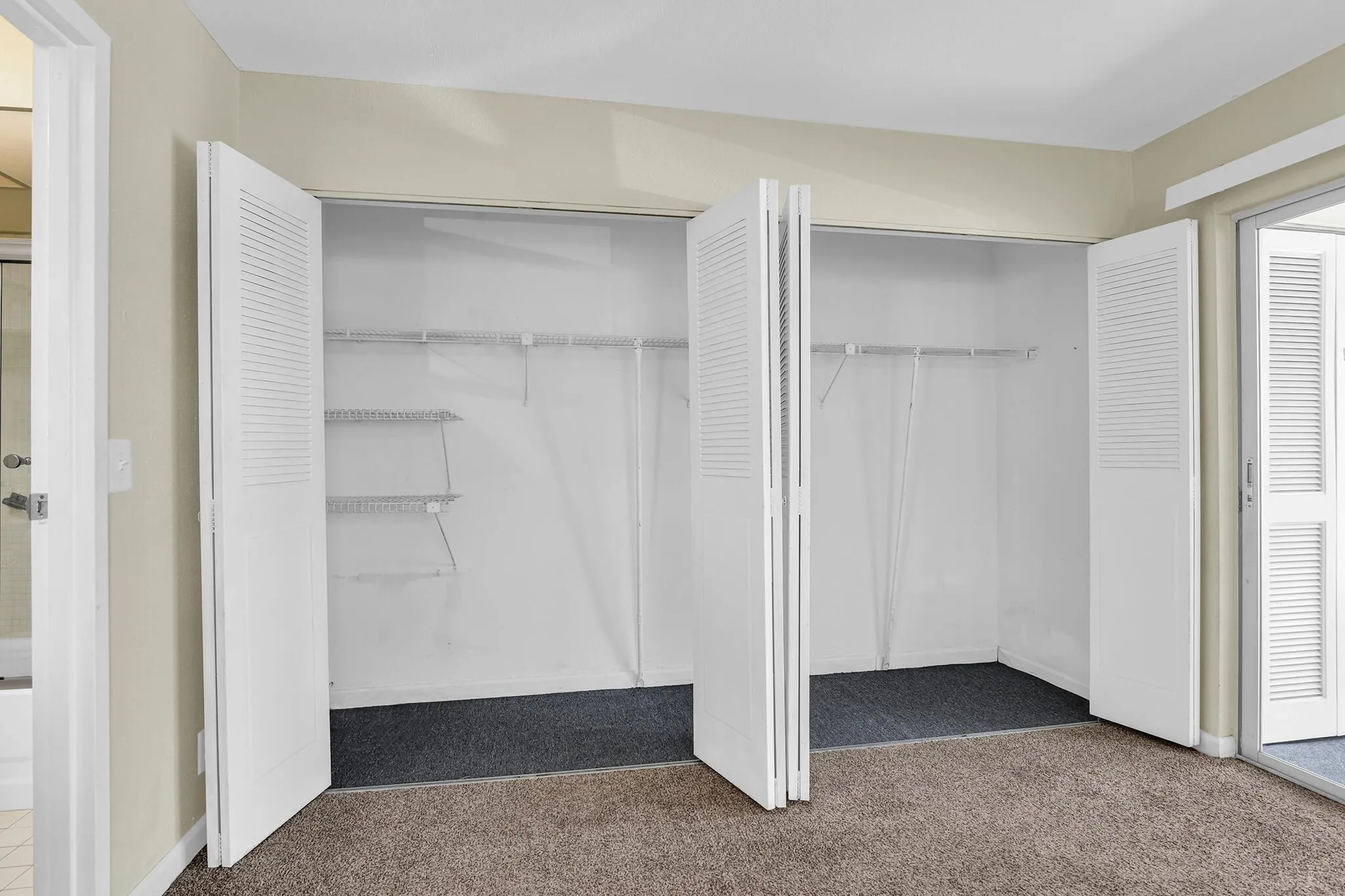 Dual louvered bi-fold closet doors reveal interior storage with wire shelving and hanging rods, set against plush carpeting