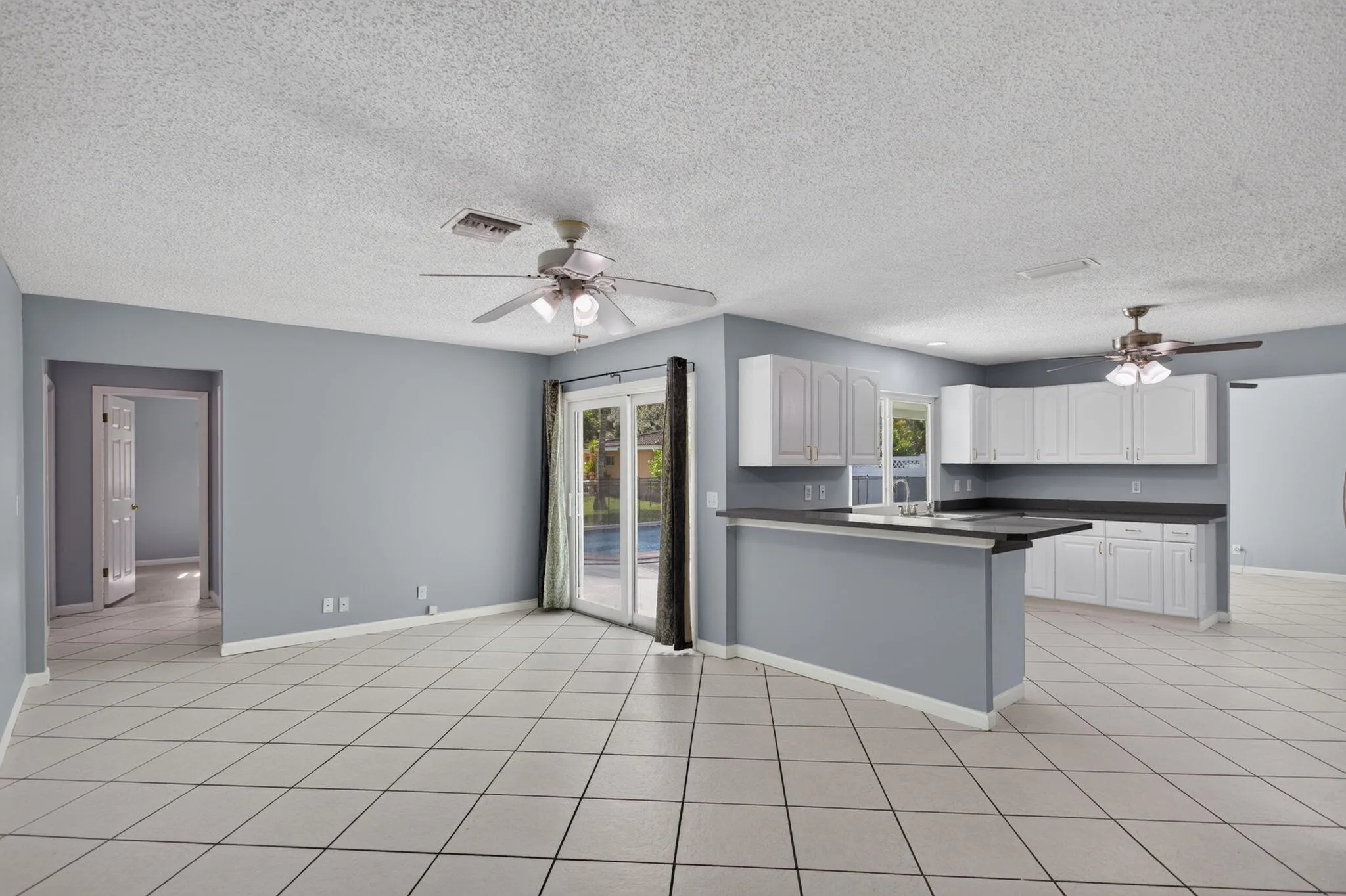 Open concept living space featuring neutral wall tones, tile flooring, two ceiling fans, a kitchen with white cabinetry and dark countertops, and sliding glass doors to the exterior
