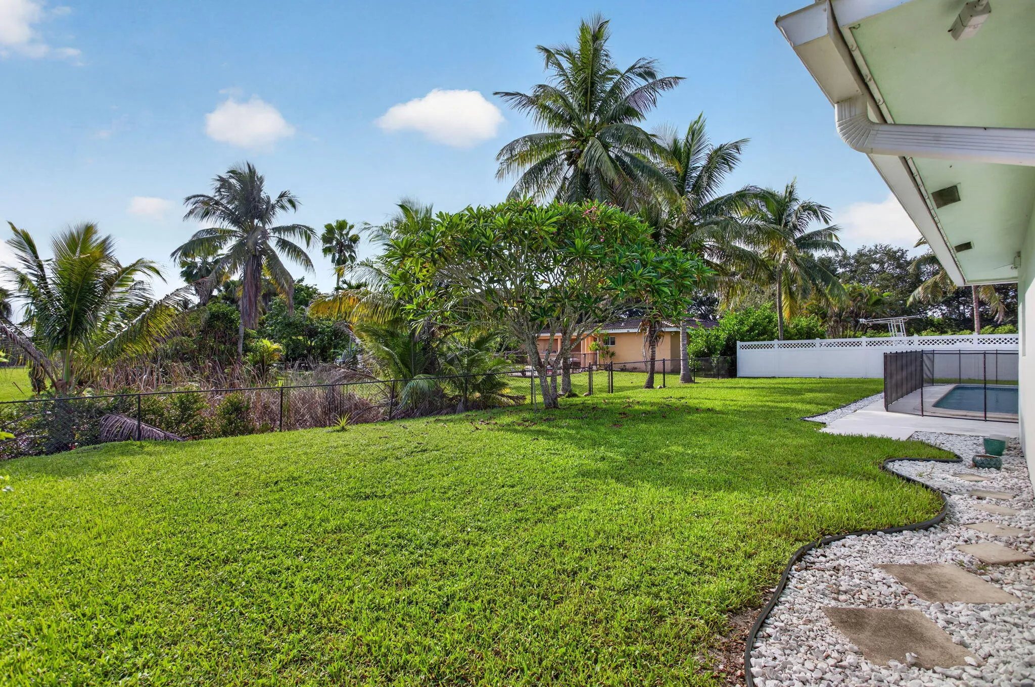 Manicured lawn, mature tropical landscaping, white privacy fence, fenced pool area, and gravel pathway with stepping stones