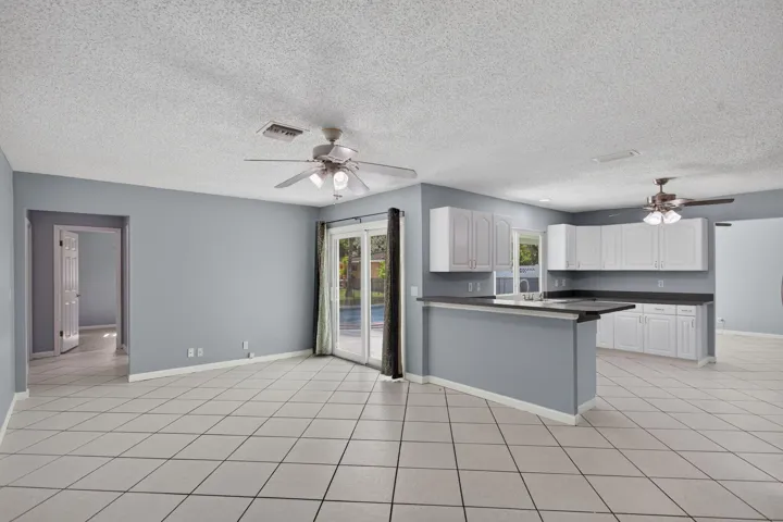 Open concept living space featuring neutral wall tones, tile flooring, two ceiling fans, a kitchen with white cabinetry and dark countertops, and sliding glass doors to the exterior