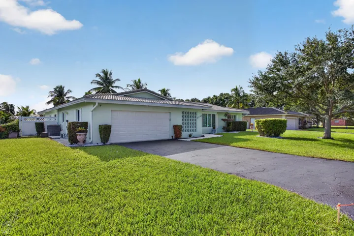 Light green exterior with a two-car garage, gray tile roof, paved driveway, and established landscaping
