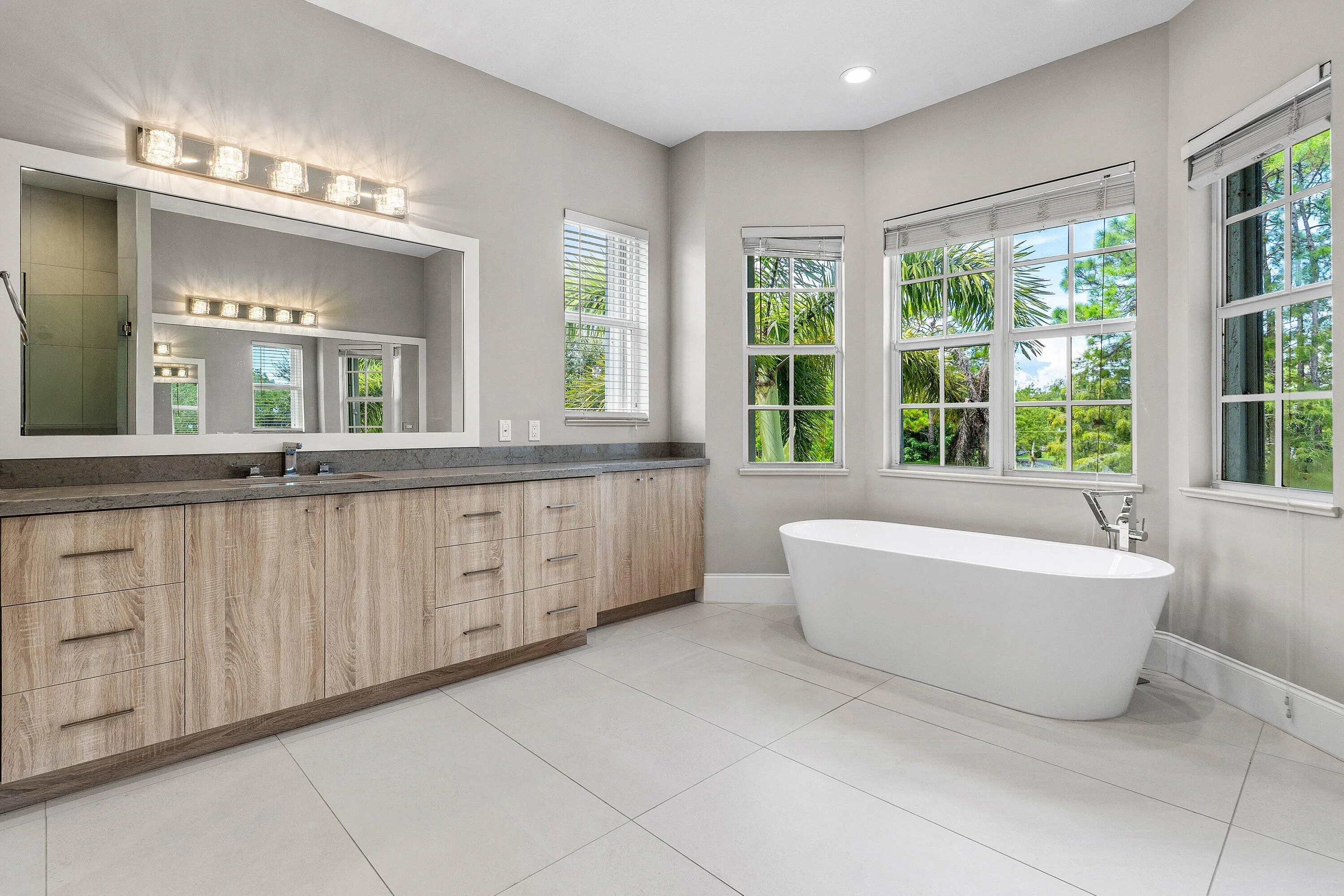 Freestanding soaking tub, dual-sink vanity with quartz countertop, wood-finish cabinetry, and large windows with privacy blinds