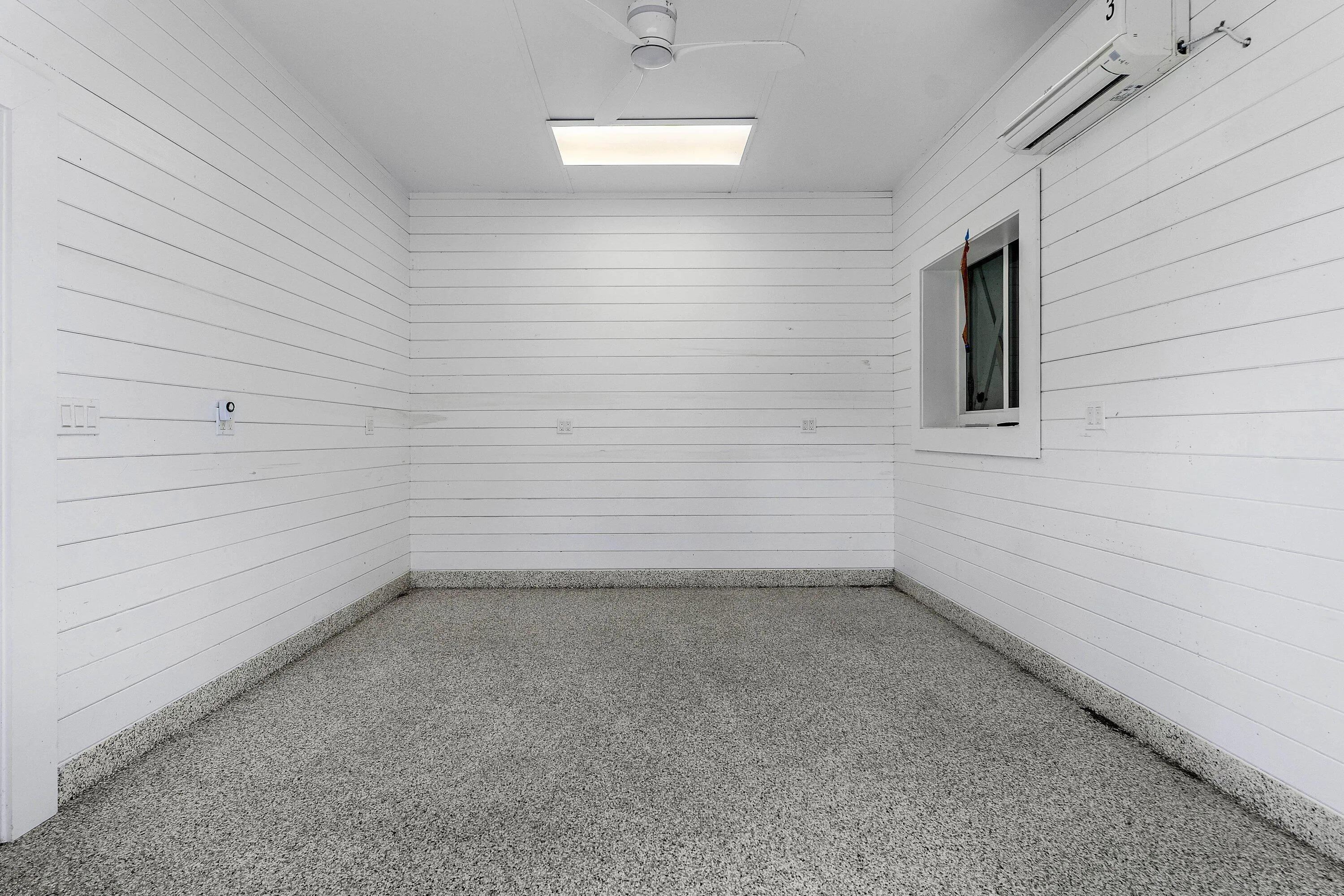 White shiplap walls, speckled epoxy flooring, ceiling fan, flush-mount lighting, and wall-mounted air conditioning unit