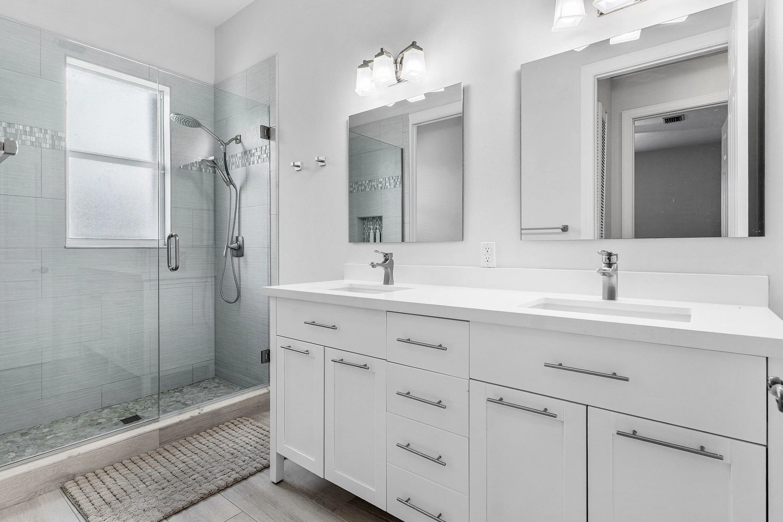 Dual vanity with white cabinetry and integrated sinks, a walk-in shower with glass enclosure, light-toned tile flooring, and wall-mounted light fixtures