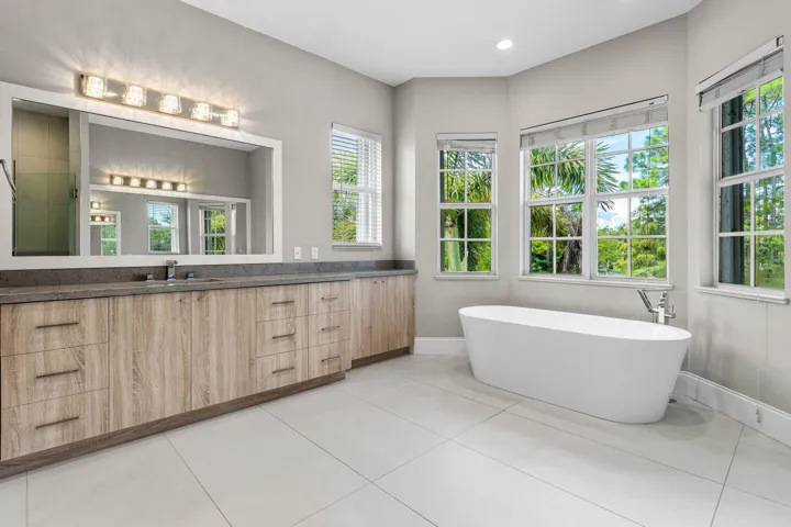 Freestanding soaking tub, dual-sink vanity with quartz countertop, wood-finish cabinetry, and large windows with privacy blinds