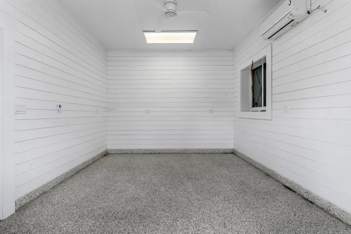 White shiplap walls, speckled epoxy flooring, ceiling fan, flush-mount lighting, and wall-mounted air conditioning unit