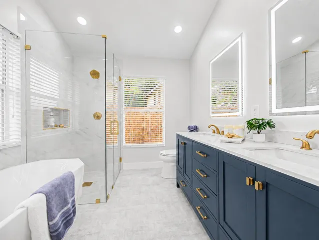 Dual vanity with matte gold fixtures and integrated lighting, glass-enclosed shower with tile surround, freestanding soaking tub, and natural light from multiple windows
