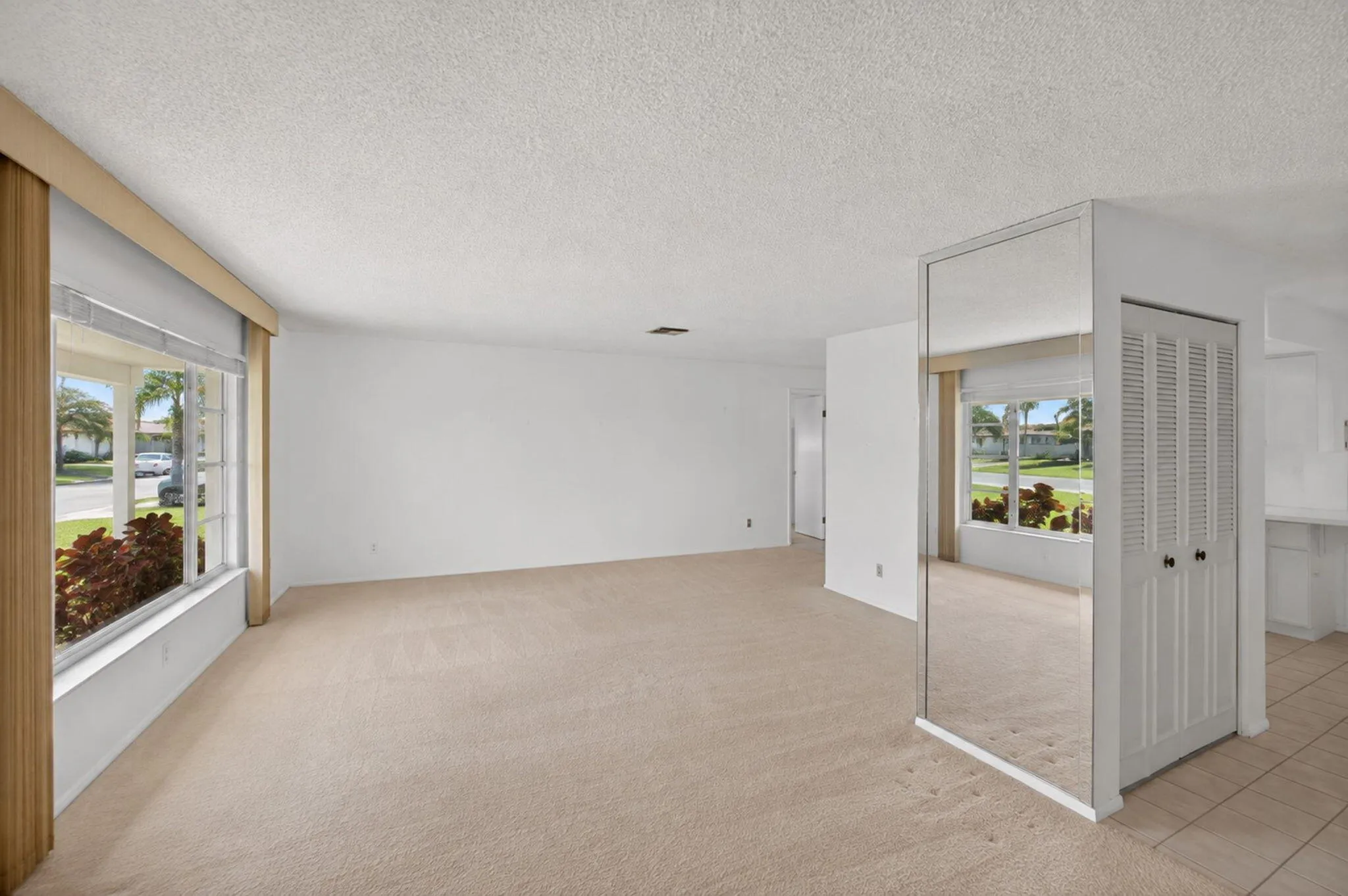 Neutral carpet flooring, white painted walls, large window with outdoor landscaping, mirrored wall accent, louvered bi-fold closet doors