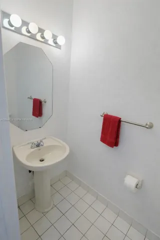 Clean half-bath with pedestal sink, octagonal mirror, bright lighting, and white tile flooring. Simple and well-maintained space.