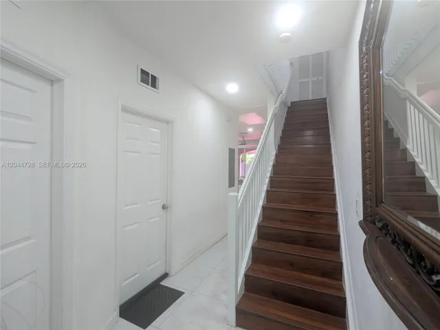 Bright interior hallway with white tile flooring and dark staircase. Clean layout with multiple doors and a large decorative mirror enhancing the space.