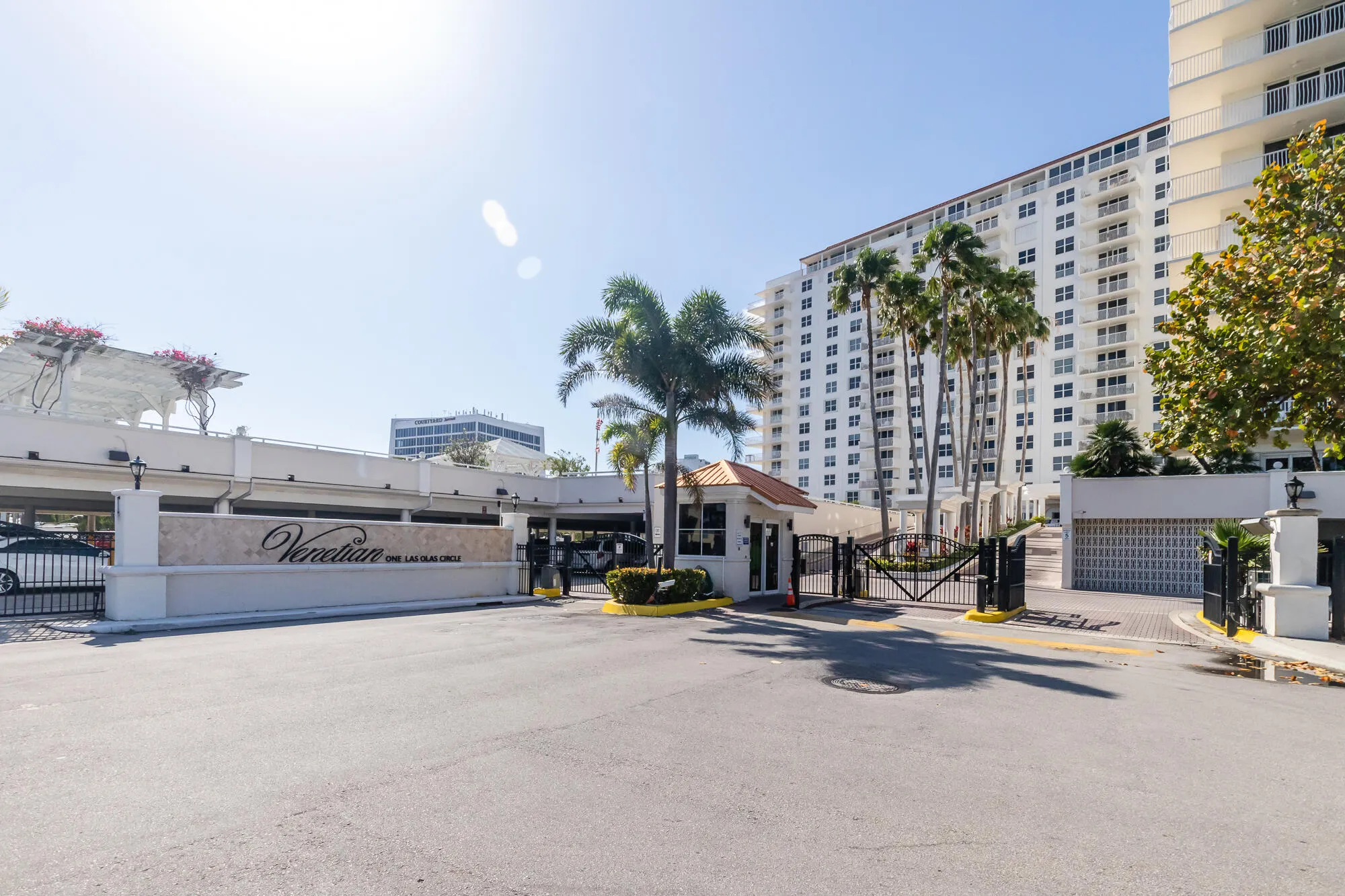 Entry gate with decorative fencing, mature palm trees, multi-story white building, and integrated security booth