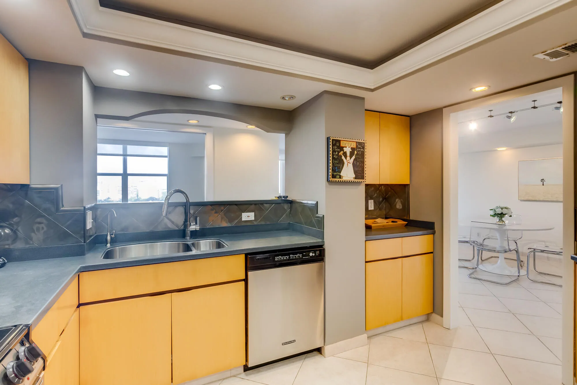 Kitchen featuring a double stainless steel sink, light wood-finish cabinetry, stainless steel dishwasher, tray ceiling with recessed lighting, and tile flooring