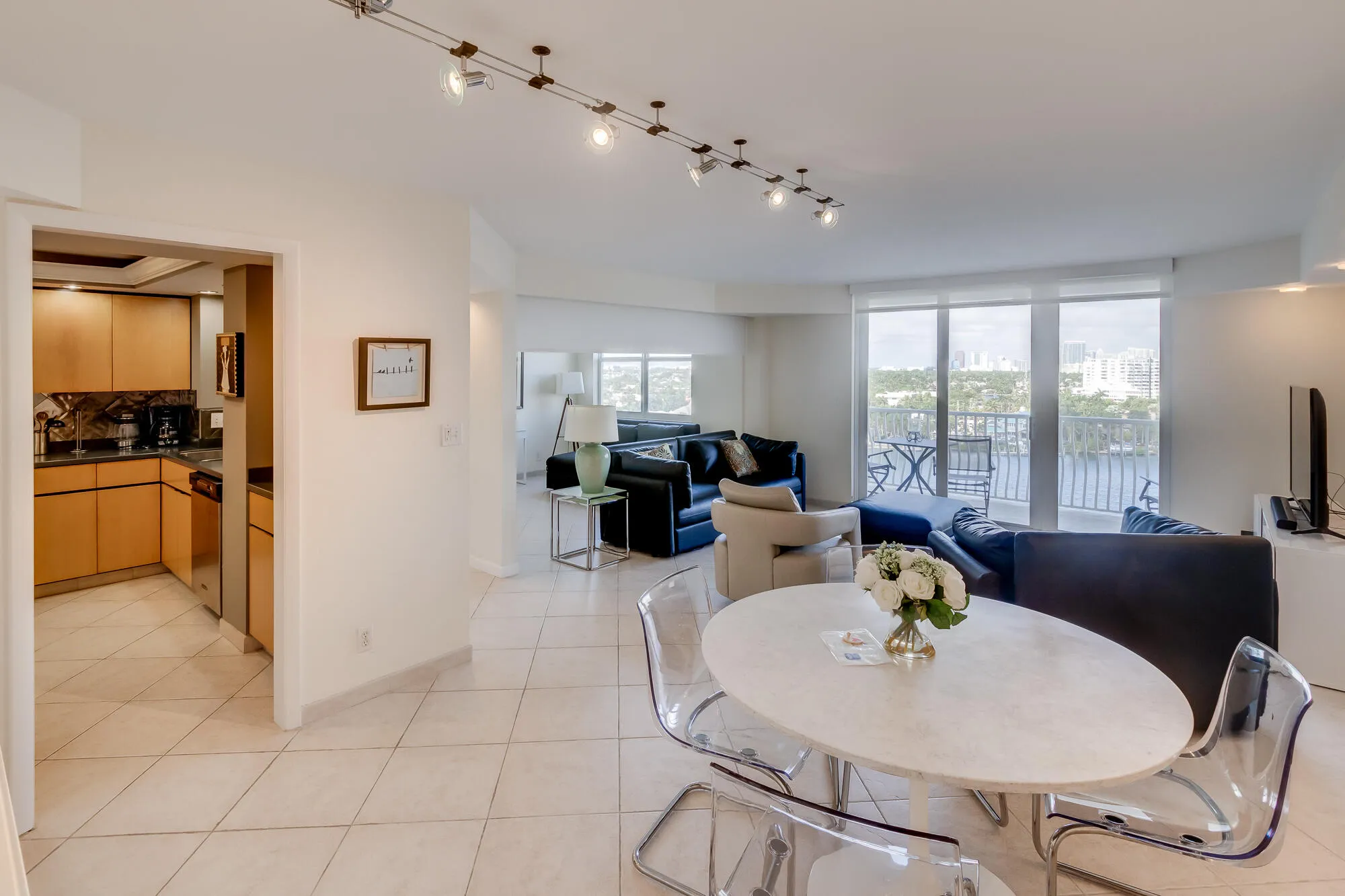 White tile flooring throughout, kitchen with wood-finish cabinetry and dark countertops, track lighting fixture, glass sliding door leading to an outdoor balcony with railing