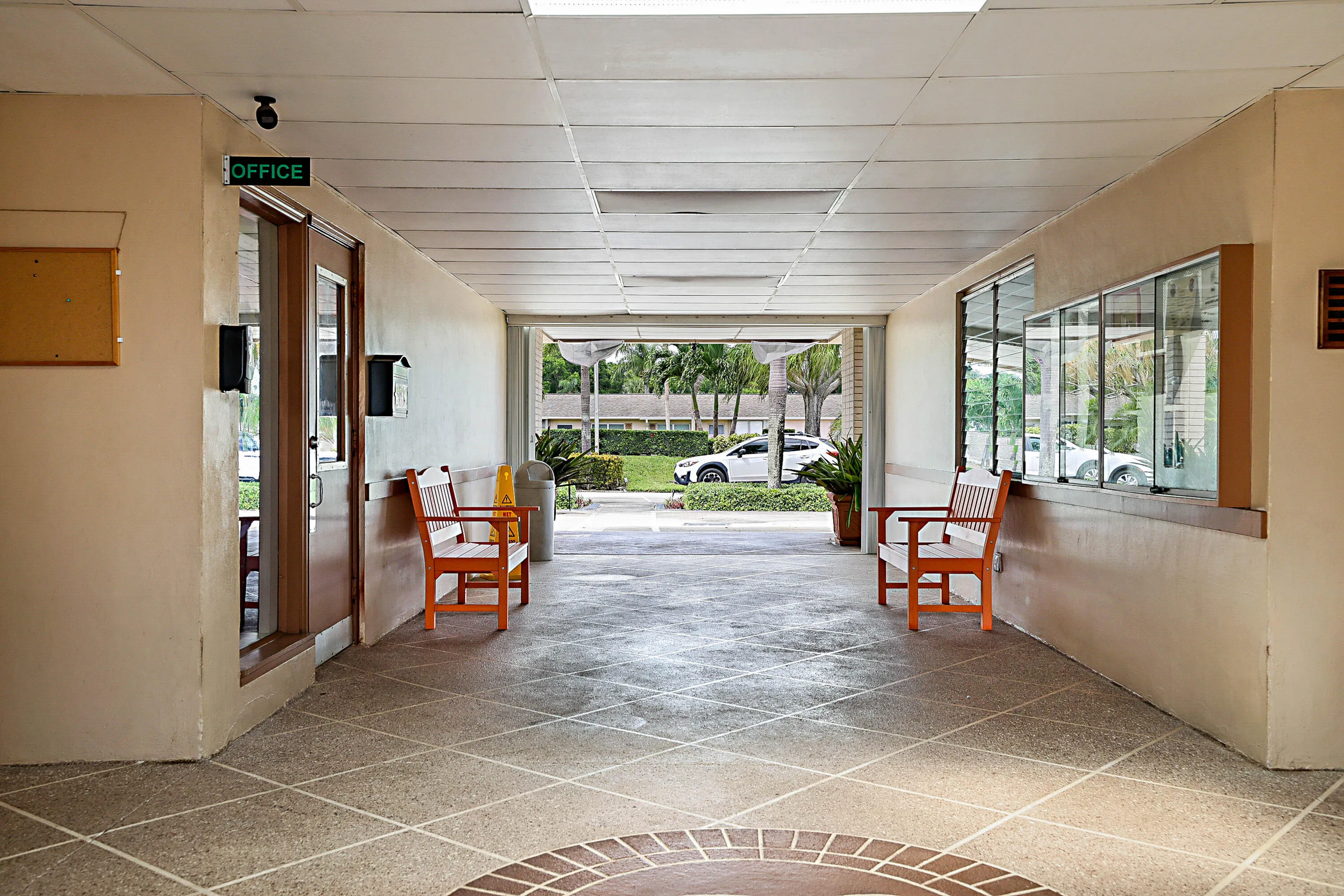 Covered walkway with a stone-finish tile floor featuring an inlaid brick pattern, providing access to managment office with a glass-paneled door