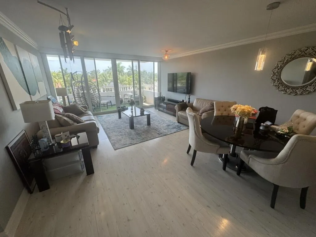 Light wood-finish flooring extending throughout, spacious living area with crown molding, glass sliding doors leading to an outdoor balcony, built-in recessed lighting, and a modern dining area with an oval-shaped table