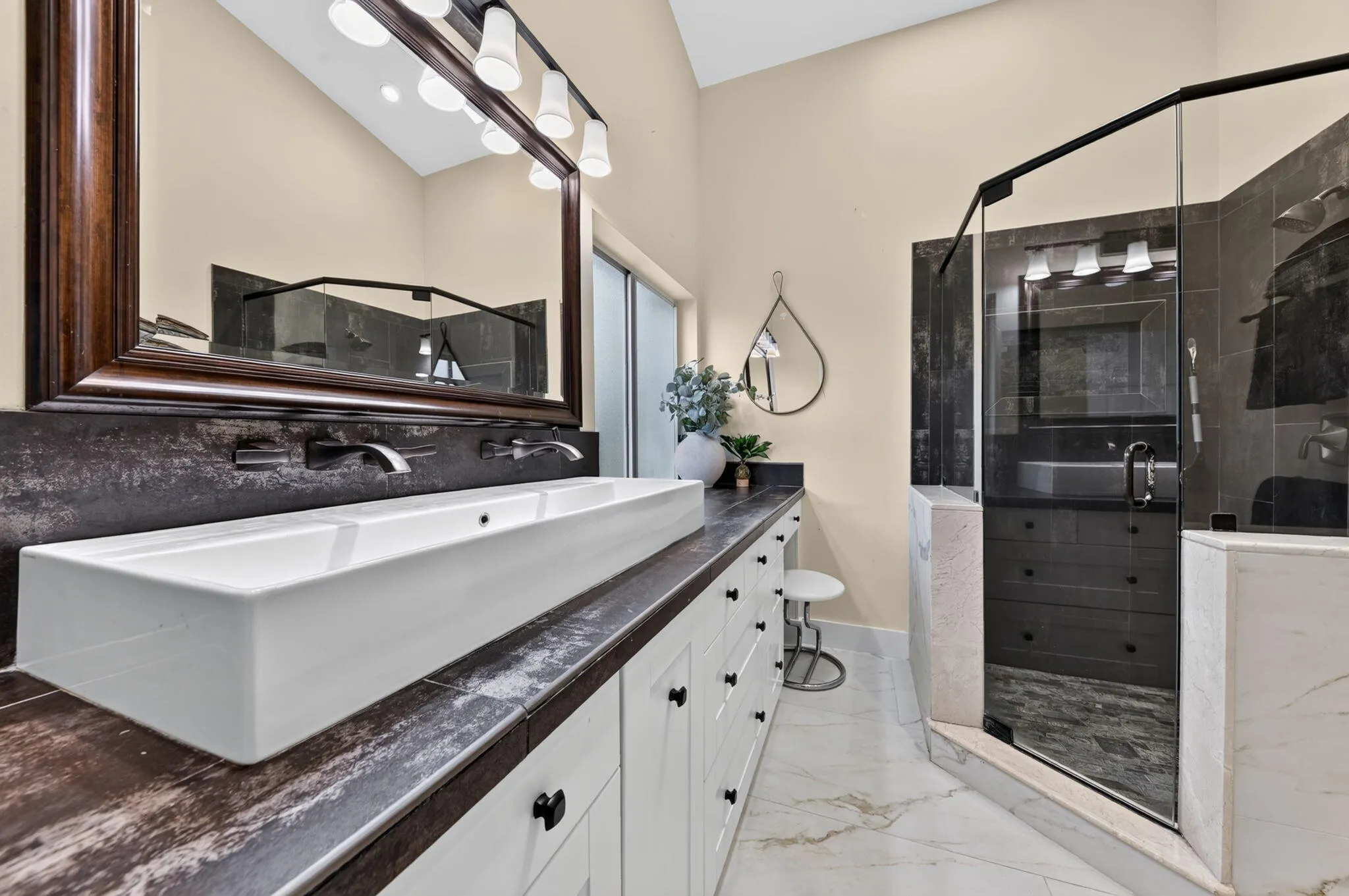Vaulted ceiling, dual wall-mounted faucets, trough sink, white cabinetry with dark hardware, and a glass-enclosed shower with dark tile