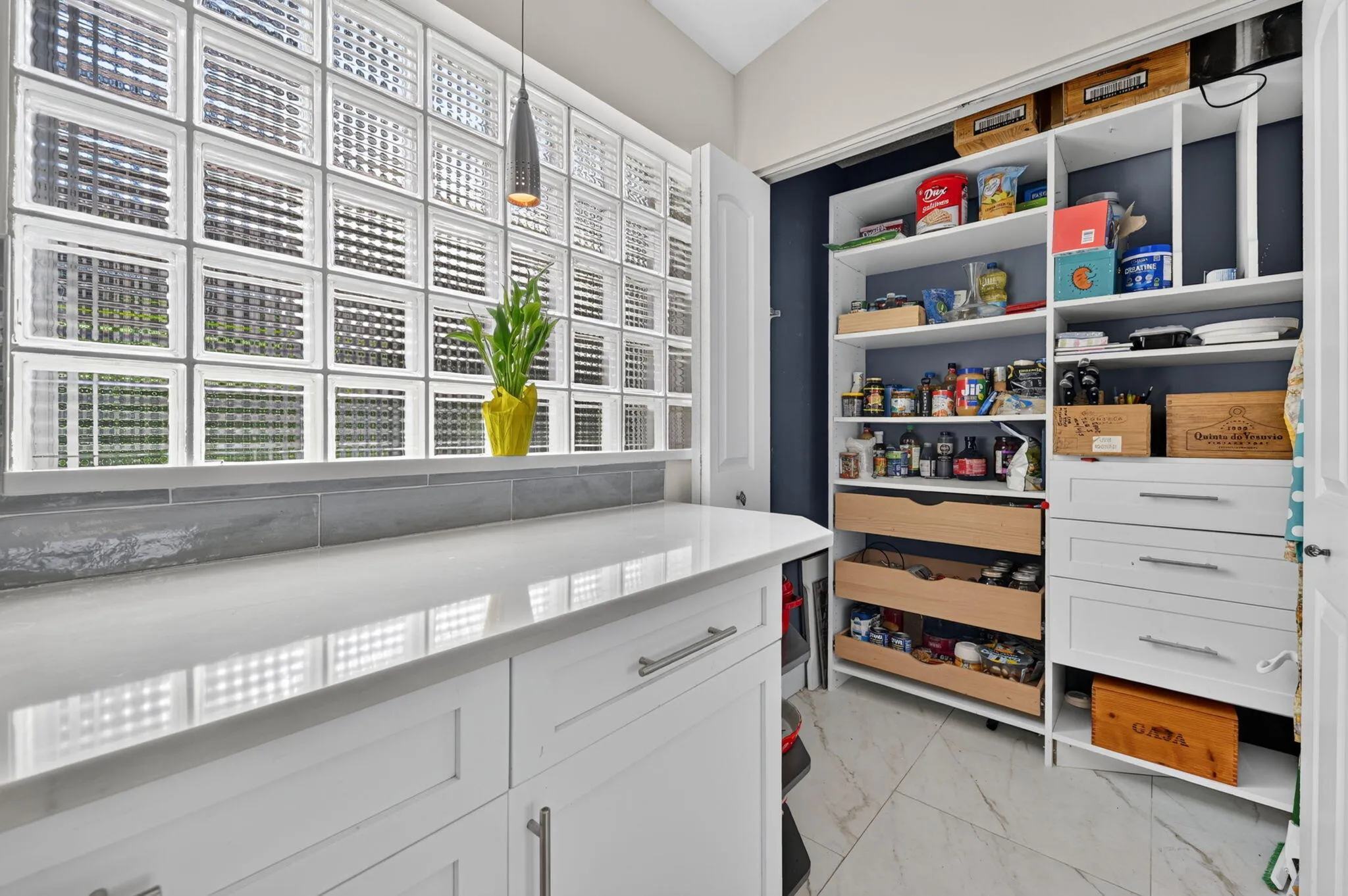 Built-in pantry storage with shelving and drawers, white cabinetry with polished hardware, quartz light-toned countertop, and a glass block window