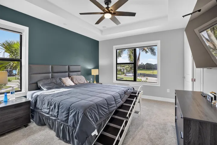 Primary bedroom suite featuring a tufted upholstered headboard, ceiling fan with brushed metal finish, and expansive windows overlooking manicured grounds