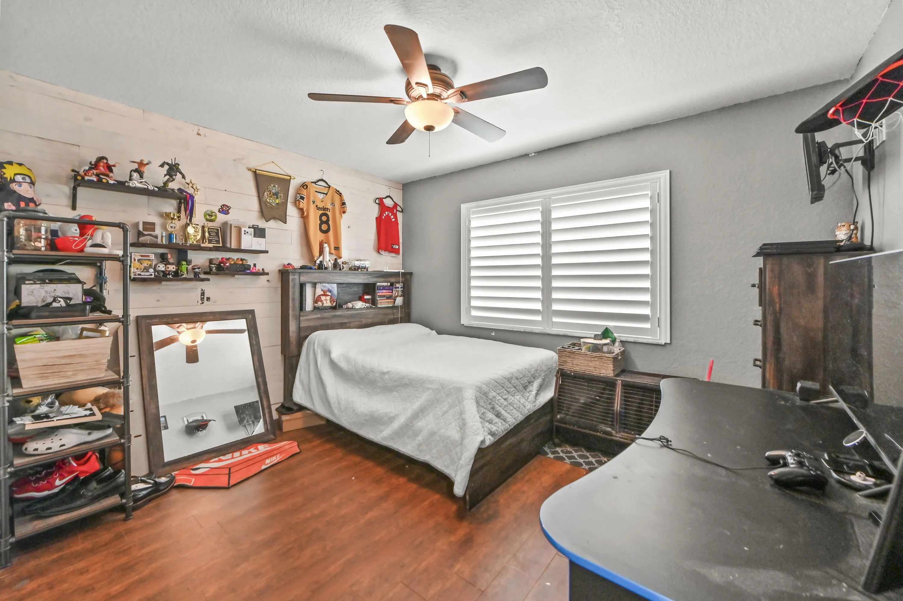 Wood-finish flooring, ceiling fan with integrated lighting, window with plantation shutters, accent wall with horizontal paneling