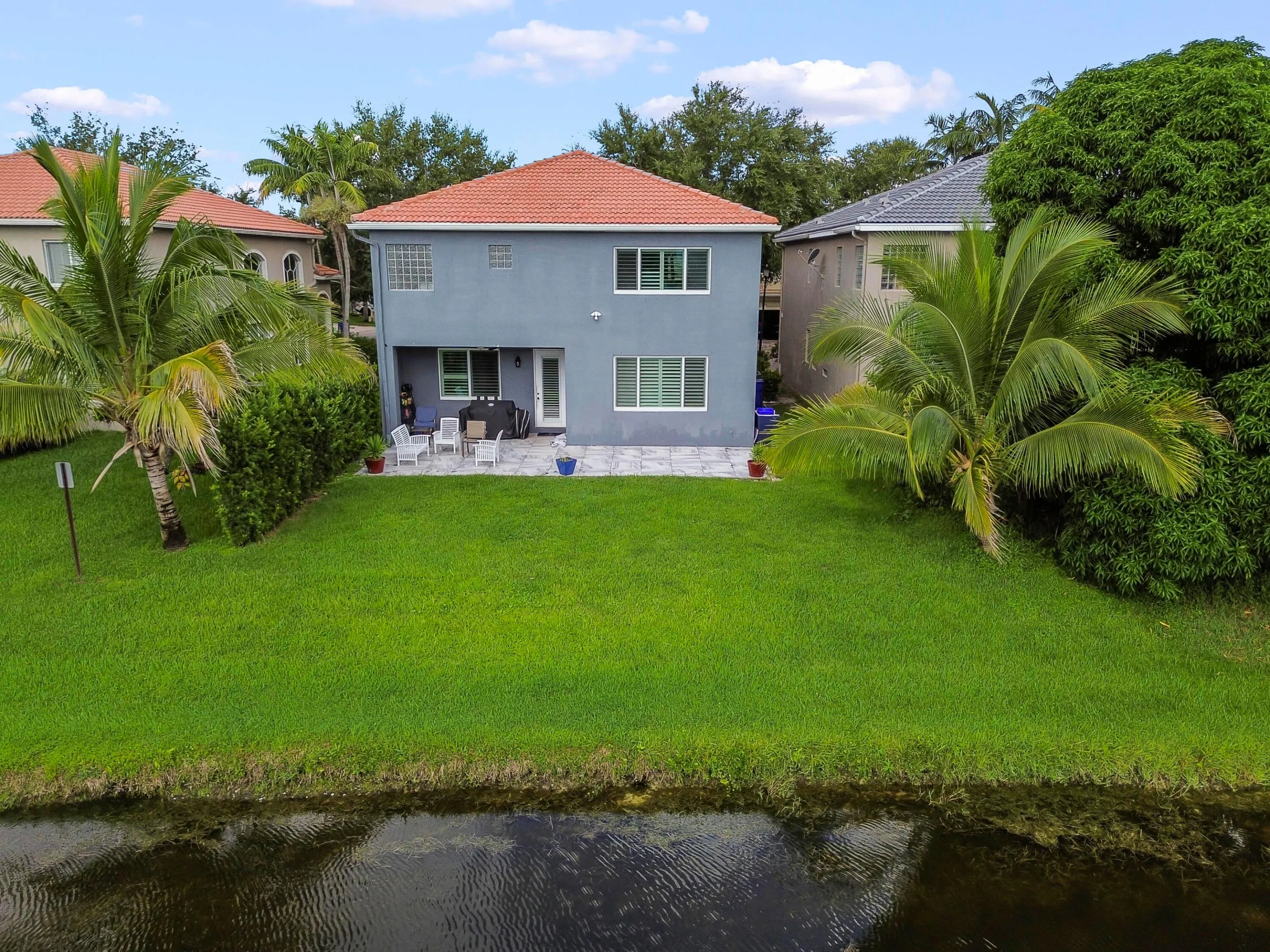 Rear exterior with a red barrel tile roof, gray stucco finish, and a ground-level patio featuring a paved surface and white window shutters