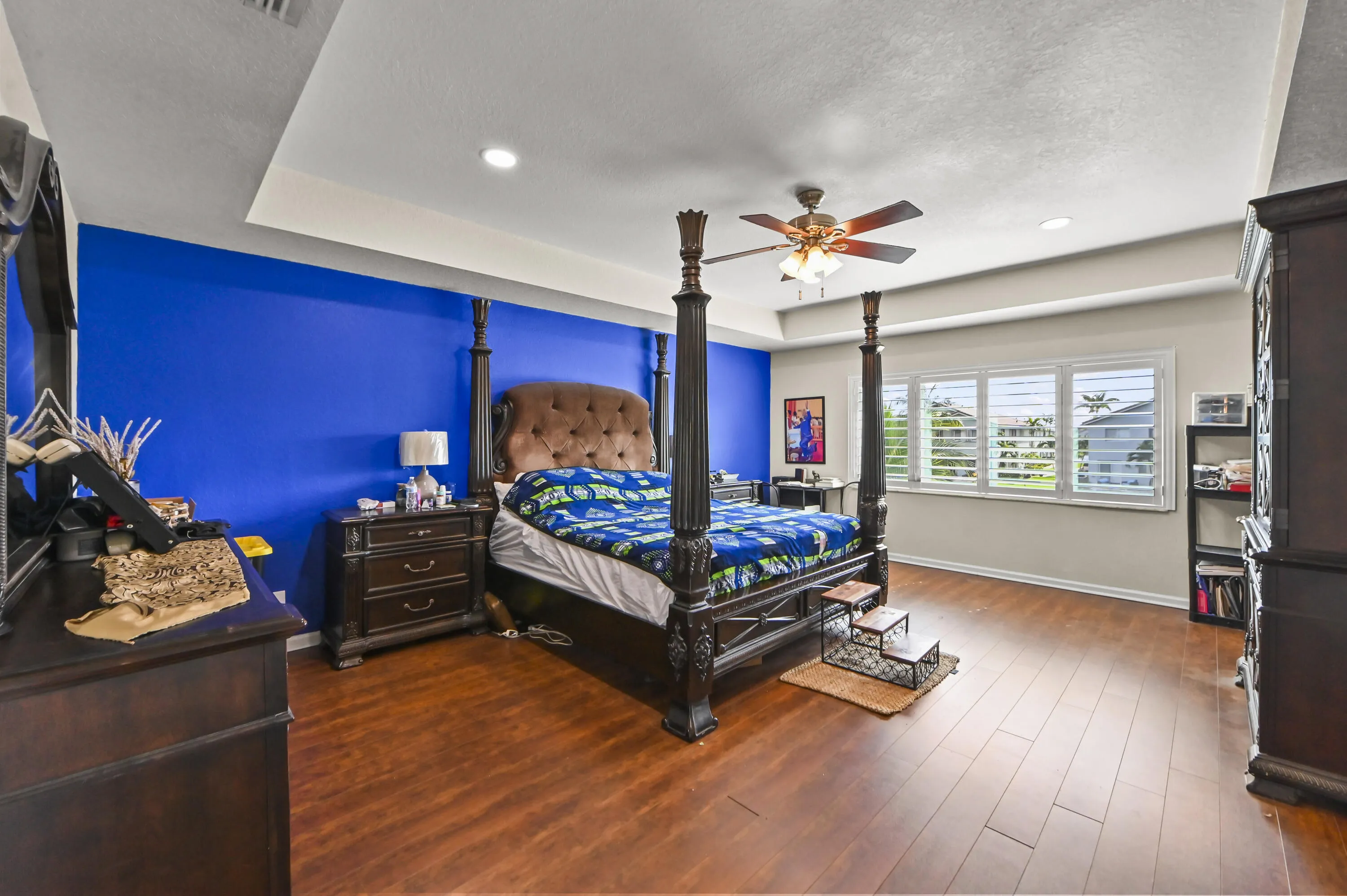 Tray ceiling with recessed lighting, wood-finish flooring, ceiling fan with integrated light, multiple windows with plantation shutters, and a distinctive accent wall