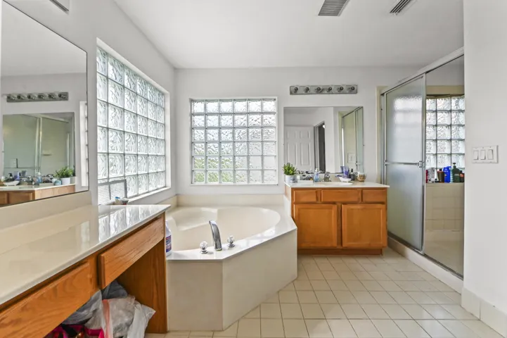 Corner soaking tub with deck-mounted faucet, dual glass block windows, ceramic tile flooring, twin vanity with wood cabinetry, and a glass-enclosed shower