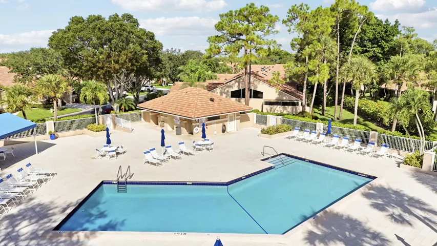 Geometric-shaped swimming pool with an expansive paver deck, surrounded by mature landscaping and a tiled-roof cabana building