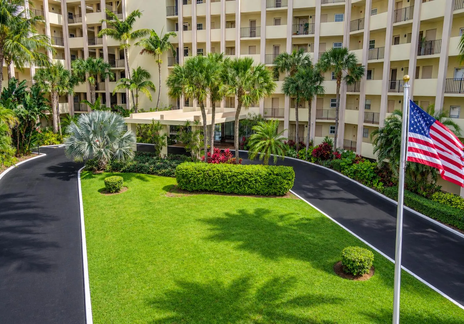 Multi-story building exterior with individual balconies, lush landscaping, mature palm trees, and a paved driveway