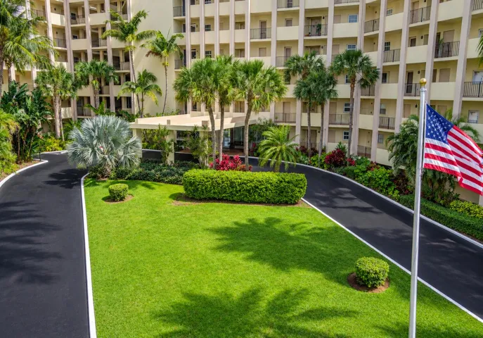 Multi-story building exterior with individual balconies, lush landscaping, mature palm trees, and a paved driveway