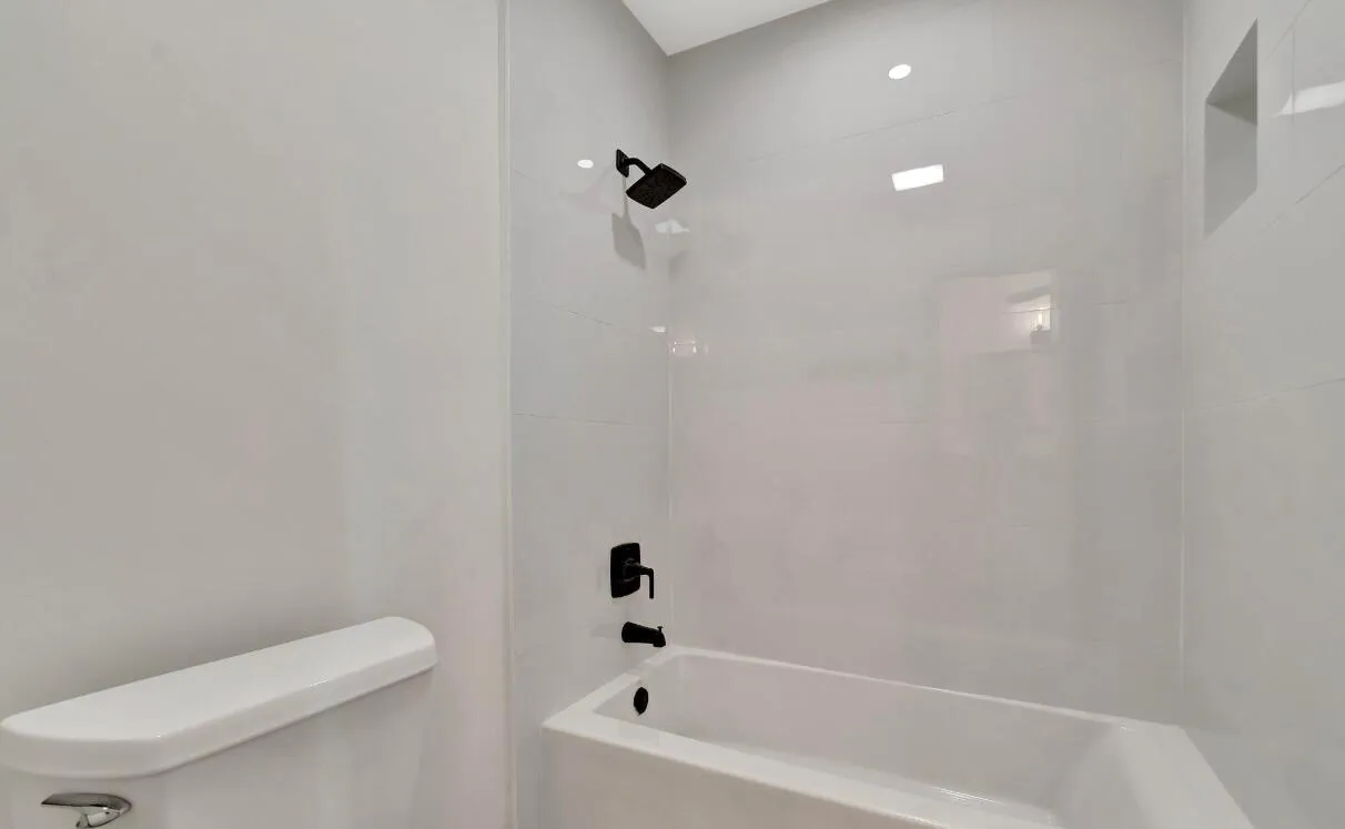Tub and shower combination with matte black fixtures, recessed shelf, and light-toned tile surround
