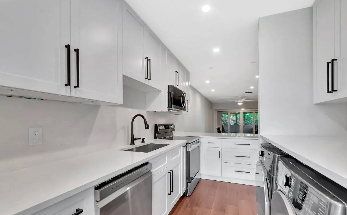 Modern galley kitchen featuring white cabinetry with black hardware, quartz countertops, stainless steel appliances including a built-in microwave and dishwasher, a gooseneck faucet with pull-down spray, recessed lighting, and warm wood-finish flooring