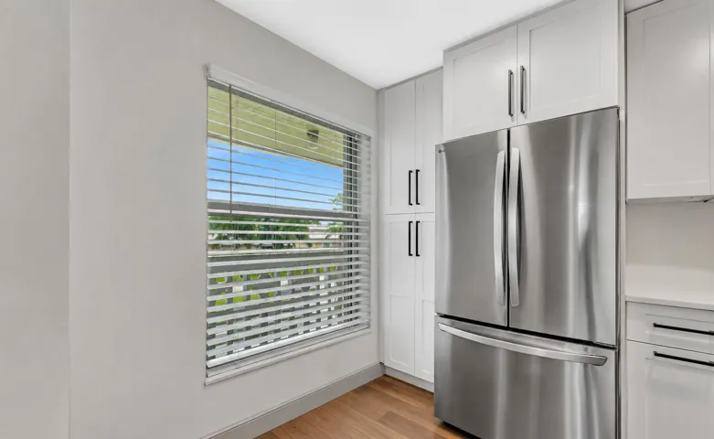 Stainless steel French door refrigerator, white shaker-style cabinetry with matte black hardware, wood-finish flooring, and window with horizontal blinds